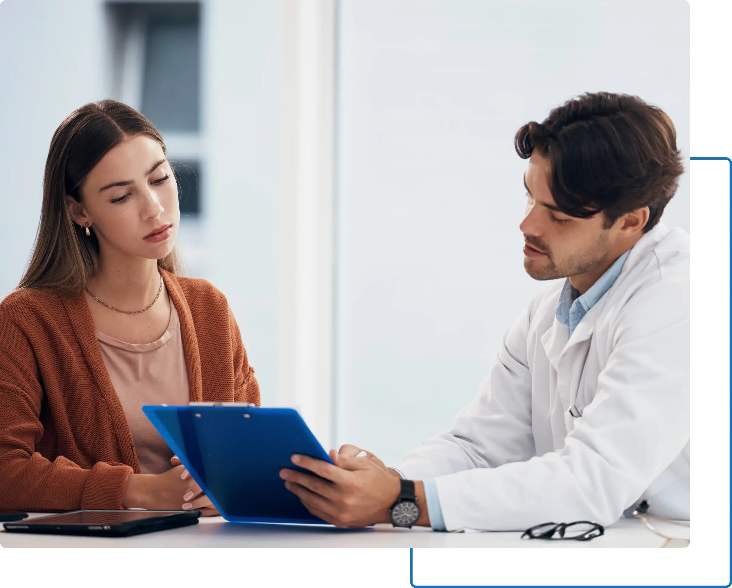 A doctor is explaining something to a woman sitting across the table, who is looking at a clipboard with a serious expression.